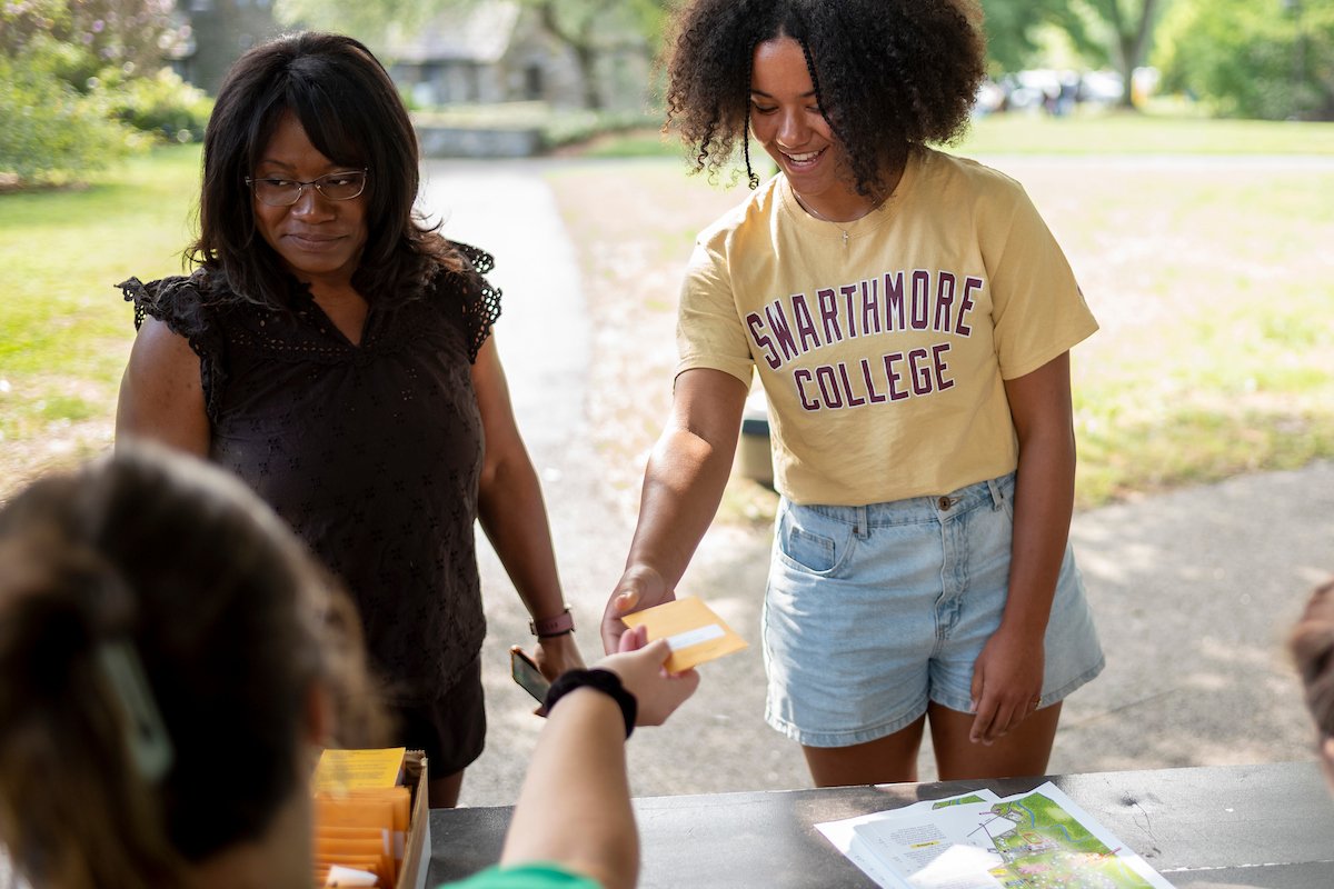 Two Black women, one older and one college-age, stand in front of a check-in table. The younger one wears a shirt that says "Swarthmore College"