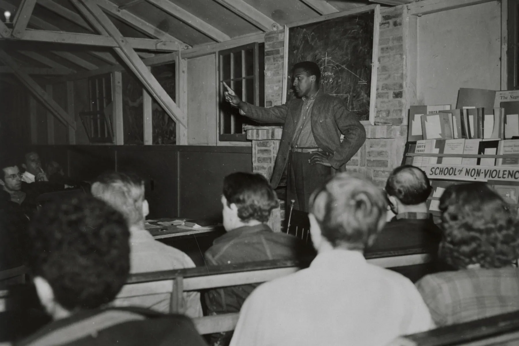 Bayard Rustin is speaking to a seated group. He is a Black man standing in front of a chalkboard. He has one arm raised in a pointing gesture. The group participants are mostly facing away from the camera, but appear to be mainly white men and one women. There is a rack of handouts with a sign that says "school of non-violence."