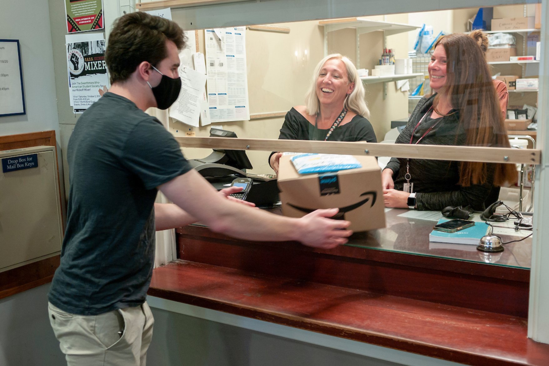 Man in grey shirt standing at the post office window, handing a package to two smiling women behind the counter