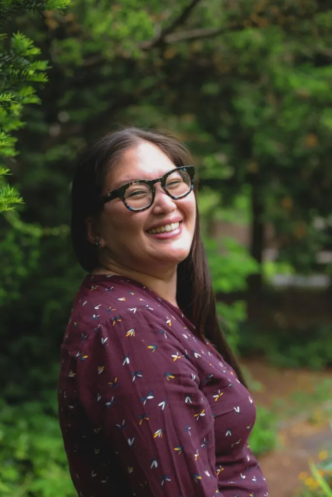 woman with long brunette hair smiles at the camera in front of a tree