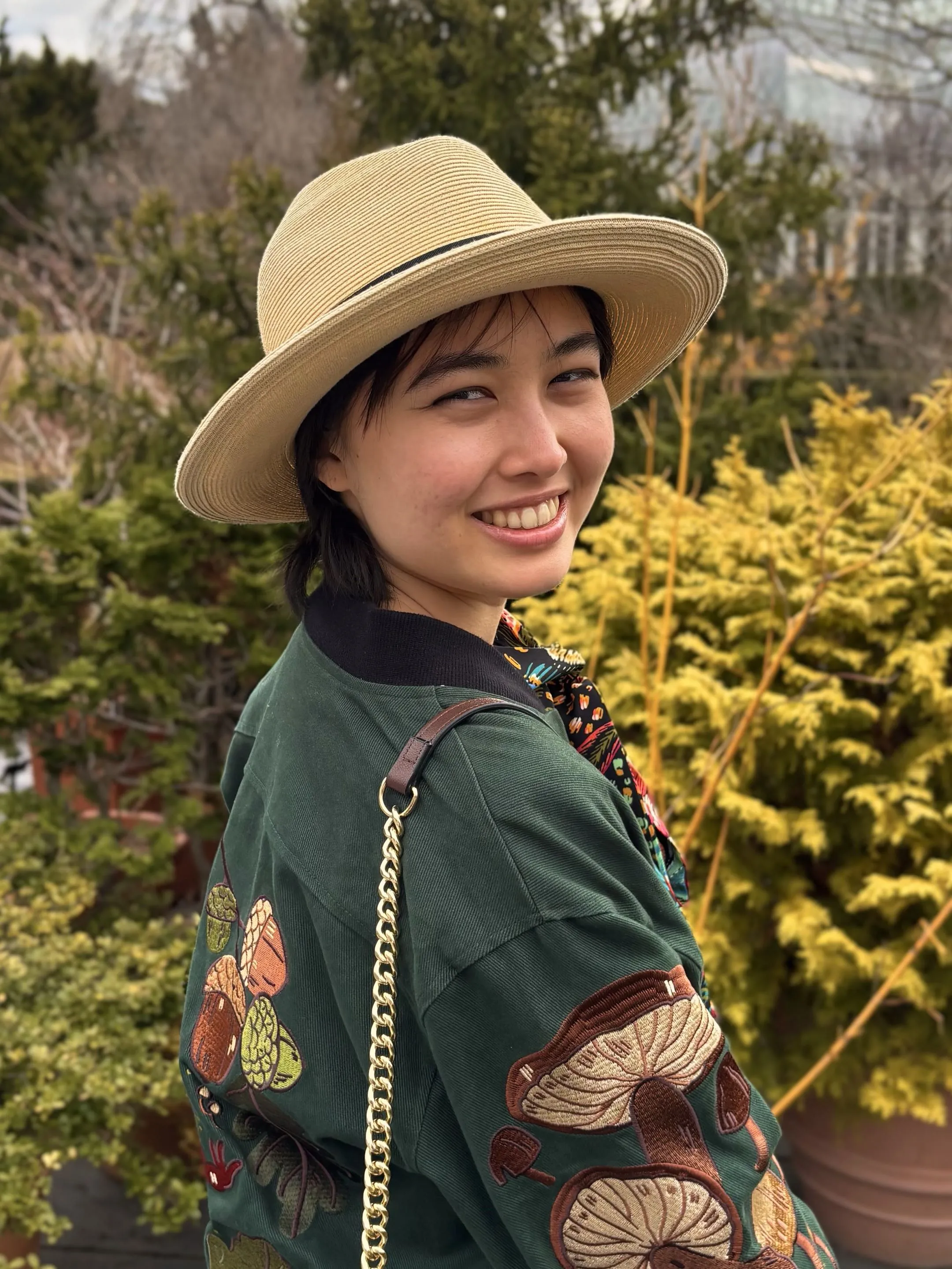 Josie Low, wearing a straw brimmed hat and jacket with fungi embroidery walking near blooming flowers