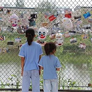 Two individuals look at body of water through a chain link face with masks