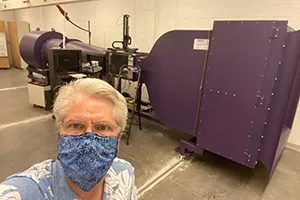 Man in mask stands indoors with wind tunnel machine in background