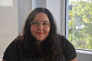 Woman with glasses smiling in front of a window with a tree outside.