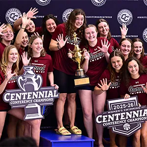 Women's swimming team celebrates conference title with group photo on podium and holding trophy while holding up five fingers.